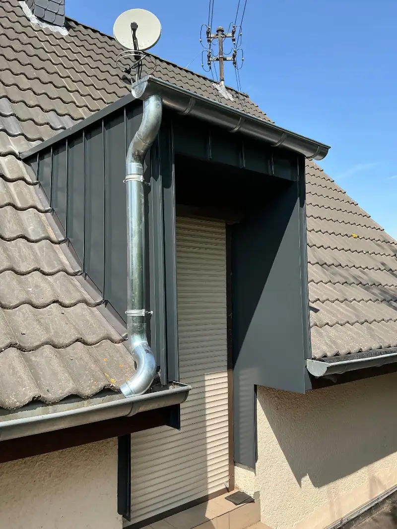 Gaube mit dunkler Metallverkleidung, silbernem Fallrohr und Dachrinne auf einem geneigten Dach
