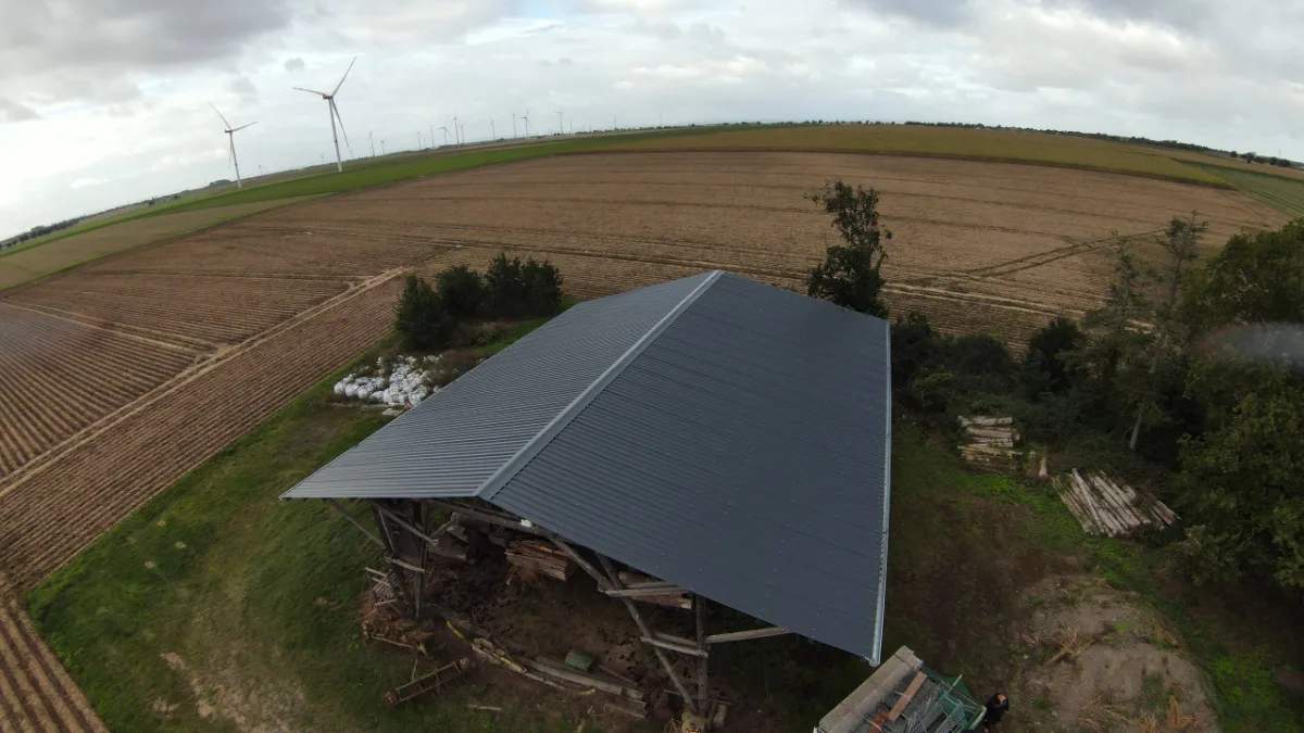 Drohnenaufnahme einer Scheune mit neuem dunklem Blechdach zwischen Feldern, im Hintergrund Windräder